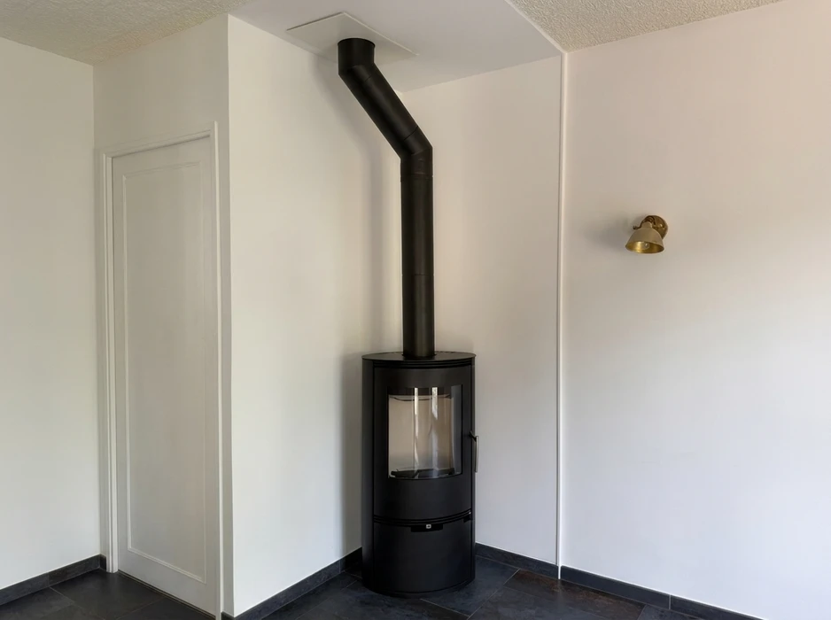 Intérieur minimaliste blanc avec poêle à bois cylindrique noir sur socle surélevé, vitre frontale panoramique, conduit de fumée noir mat coudé traversant le plafond